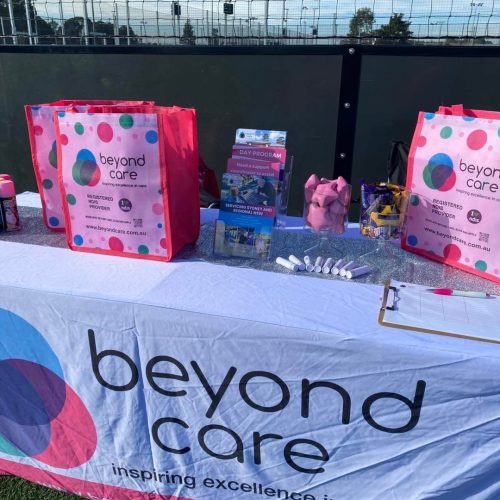 A table covered with a “beyond care” cloth displays pink gift bags, brochures, spray paint cans, and a clipboard set up for a disability-friendly community event at an outdoor gathering.