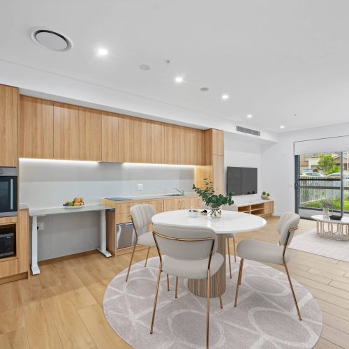 Modern open-plan kitchen and dining area with wooden cabinetry, round dining table with four chairs, and a living space featuring a sofa and large window.