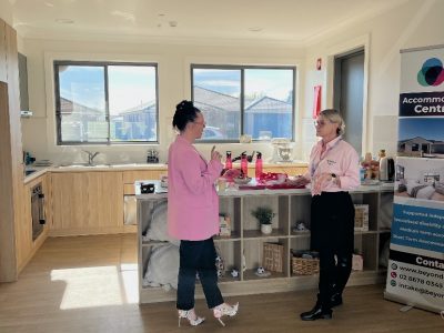 Two women stand and talk in a bright kitchen area next to a promotional banner for accommodation services.