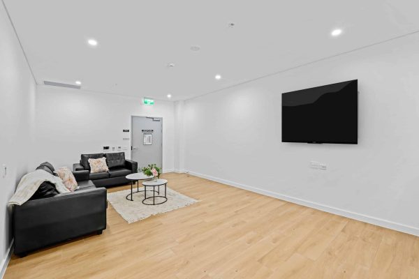 Merrylands SDA 4 Modern living room with light wood flooring, a black leather sofa, two small round tables, a rug, and a wall-mounted flat-screen TV. Walls and ceiling are white.