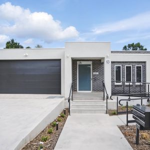 Modern single-story house with a flat roof, black garage door, brick and white exterior, front steps, ramp, and a mailbox in the foreground—perfect as Specialist Disability Accommodation (SDA NDIS) for accessible living.