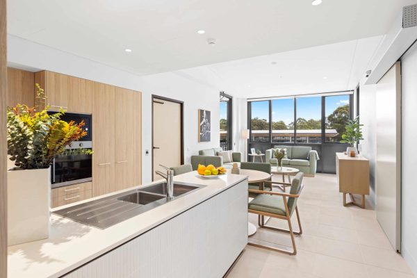 Lidcombe SDA 7 Modern open-plan kitchen and living area with wood accents, round dining table, green chairs, large windows, and sunlight streaming in.