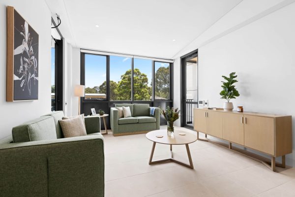 Lidcombe SDA 5 Modern living room with green sofas, a round coffee table, sideboard, large windows, and potted plants, with natural light illuminating the space.