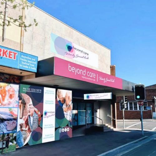 A building facade with a pink sign reading "Beyond Care" and "Nursing & Specialist Health." A contact number and various images are displayed on the storefront.