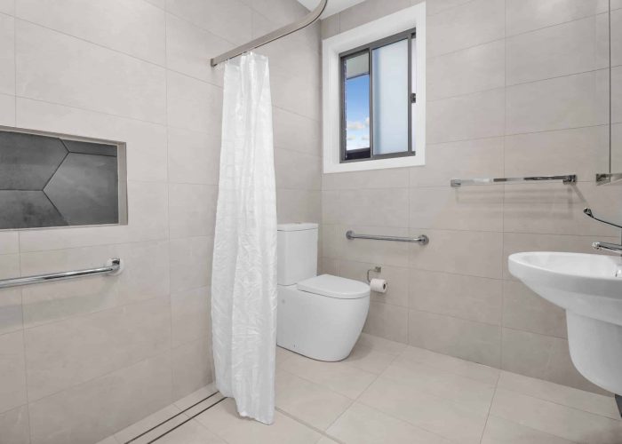 CHESTERHILL 2 3 Modern bathroom with beige tiles, a white toilet, sink, walk-in shower with a white curtain, grab bars, and a window.