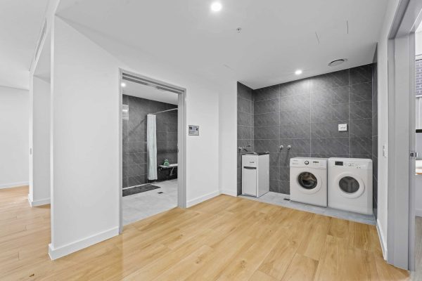 Merrylands SDA 6 Modern laundry area with a washing machine, dryer, and sink next to a bathroom; light wood flooring and grey tiled walls create a clean, minimalist look.