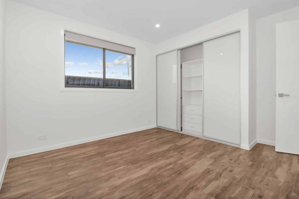 Empty room with wood flooring, a window with blinds, and a built-in closet with sliding doors and shelves. Walls and ceiling are white with a single ceiling light.