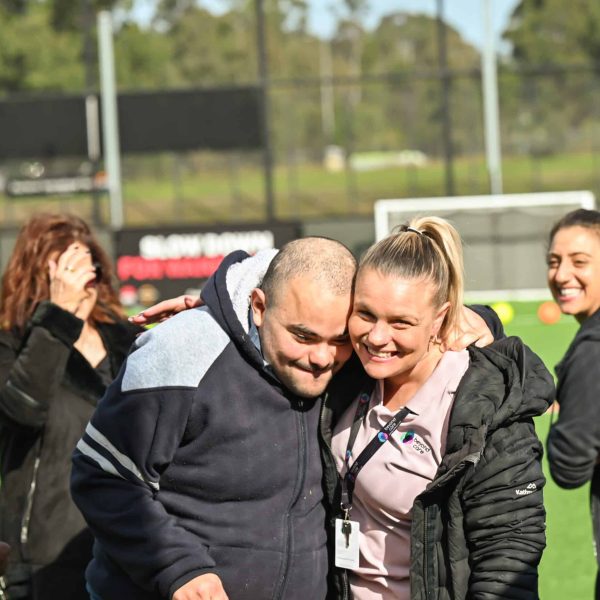 A group of people stand on a sports field, with two individuals in the foreground—participants with an NDIS provider Sydney—smiling and posing closely together for the photo.
