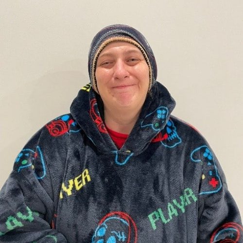 Person wearing a beanie and a dark hooded pullover with game controller patterns and the word "PLAYER" stands against a plain white background, smiling at the camera.