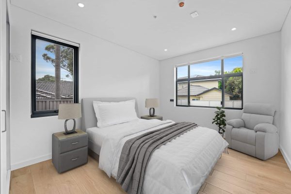 Merrylands SDA 3 A modern bedroom with a single bed, two bedside tables with lamps, a gray armchair, light wood flooring, and large windows showing an outdoor view.