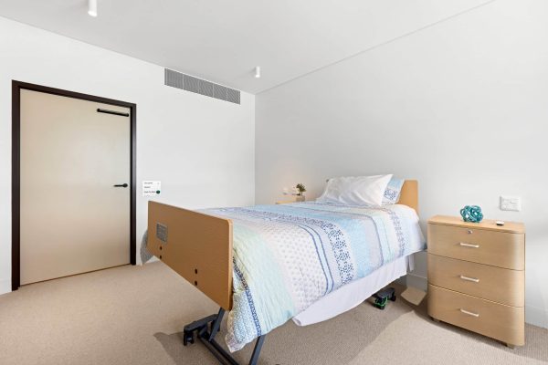 Lidcombe SDA 2 A hospital or care facility room with a single adjustable bed, a bedside table, a closed door, and neutral-colored walls and flooring.