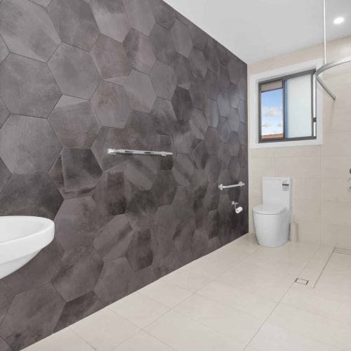Modern bathroom with large hexagonal dark tiles on one wall, beige tiles on the floor, a white sink, toilet, open shower, and a window with a view of the sky.
