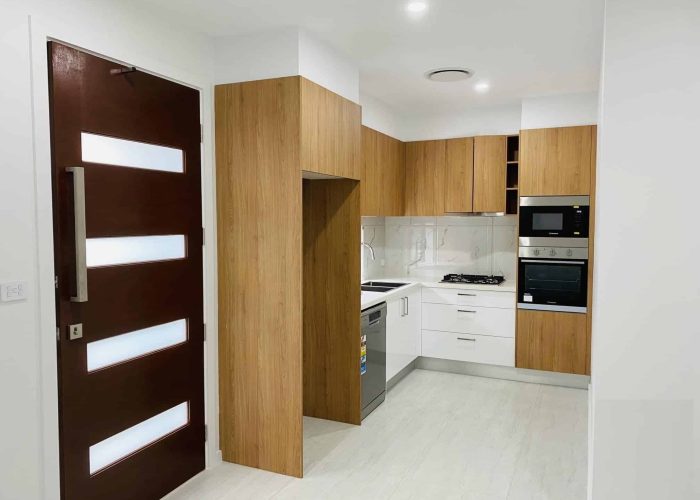 CHESTERHILL 2 5 Modern kitchen with wooden cabinets, white countertops, an integrated oven, and a gas cooktop. The floor is light-colored, and a dark wooden door with frosted panels is visible.