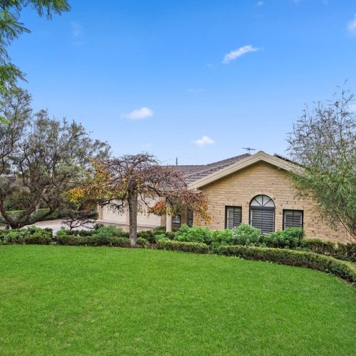 Single-story house with arched windows, tan brick exterior, and manicured green lawn surrounded by trees and hedges under a blue sky—ideal for SIL Accommodation Sydney.