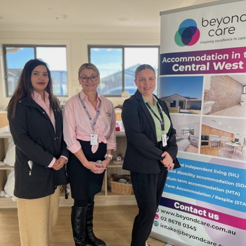 Three women stand indoors next to a Beyond Care promotional banner about accommodation services in Central West NSW.