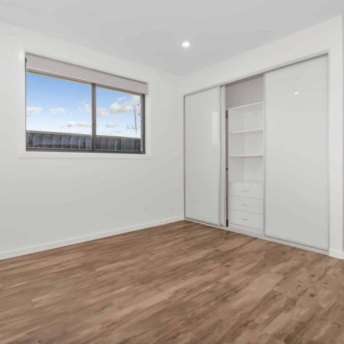 Empty room with white walls, wooden floor, a window, and a built-in closet with sliding doors. One door is open showing shelves and drawers inside. Ceiling light is on.