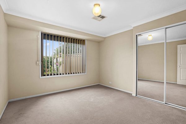 Kellyville 6 Empty beige bedroom with carpeted floor, mirrored sliding closet doors, ceiling light, vertical blinds, and a window showing greenery outside.