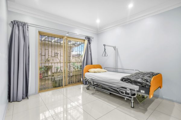 Fairfield 10 A hospital bed with orange accents is placed in a clean, white tiled room with gray curtains and large sliding glass doors leading outside.