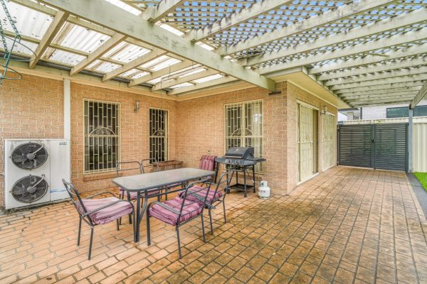 Fairfield 12 Covered patio with brick flooring, outdoor dining table with six chairs, barbecue grill, and metal lattice roof attached to a brick house.