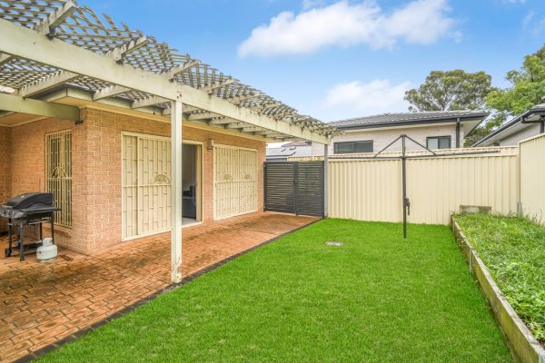 Fairfield 11 A backyard with a covered patio, brick flooring, grass lawn, metal fencing, and a barbecue grill under wooden lattice roofing.