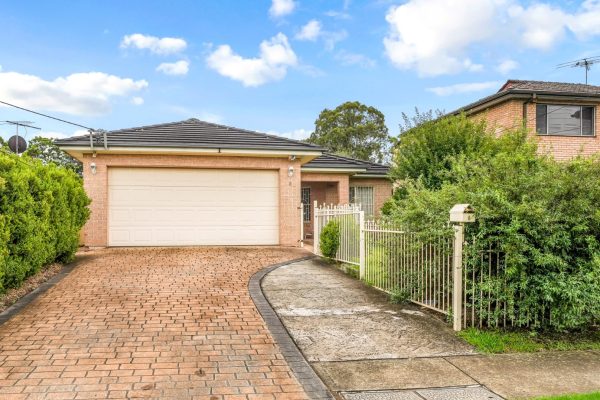 Fairfield 1 Single-story brick house with a double garage, brick driveway, and a gated side entrance, surrounded by shrubs and greenery under a partly cloudy sky.