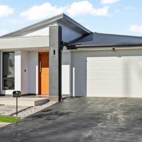 Modern single-story white house with a black roof, double garage, and wooden front door. Paved driveway and small lawn area are visible.