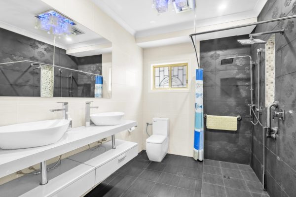 Modern bathroom with two sinks, a toilet, and a walk-in shower with a rainfall showerhead and grab bars; black and white tiles and a window above the toilet.