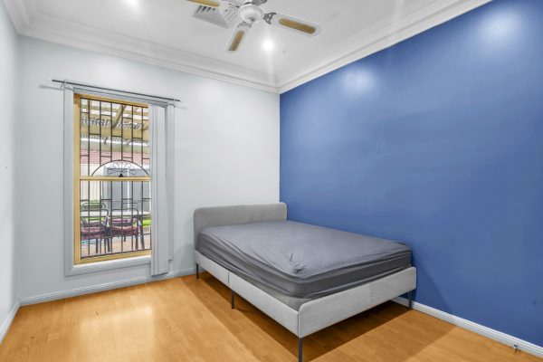 Fairfield 7 A simple bedroom with a blue accent wall, a gray bed frame with mattress, wood flooring, ceiling fan, and a window with metal bars.
