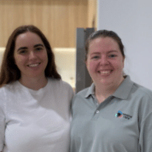 Two people are smiling at the camera. One is in a white shirt, and the other sports a light gray polo with an SDA logo. Indoors with a blurred background, they exude camaraderie and high support for their shared mission on SDA vacancy initiatives.