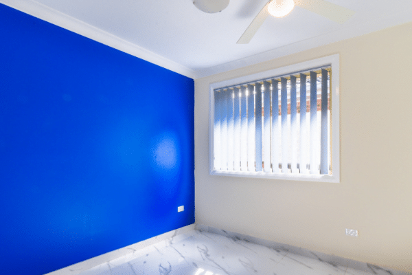 Room with a bright blue accent wall, beige walls, a window with vertical blinds, a ceiling fan, and white marble tile flooring.