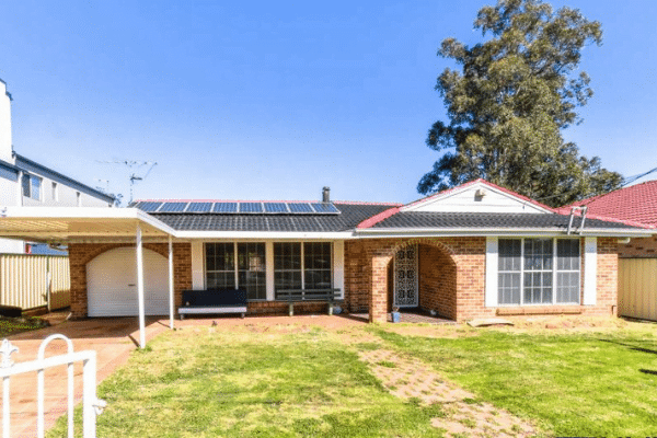 Single-story brick house with solar panels, a garage, and a front yard with grass. Large windows are present, and there is a tree on the right side.