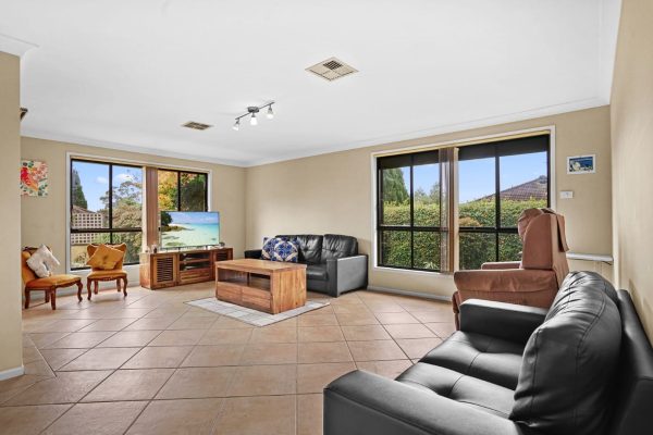Kellyville 2 Spacious living room with tiled floor, two black sofas, a brown recliner, two orange chairs, a wooden coffee table, and large windows with garden views.