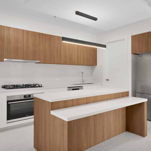 Modern kitchen with wooden cabinets, white countertops, built-in oven and gas stove, stainless steel refrigerator, and a central island with seating. Bright overhead lighting.