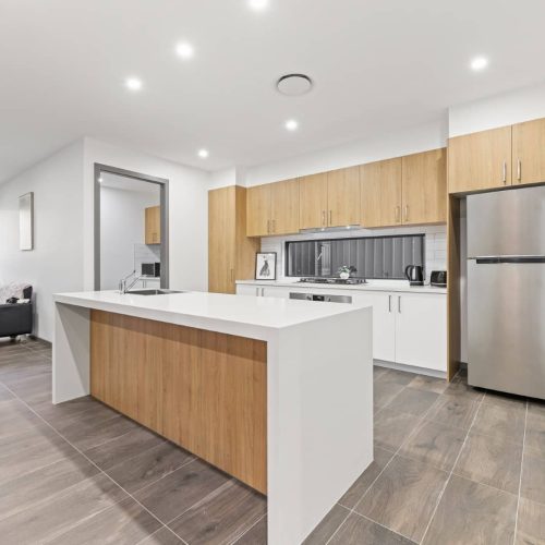 Modern kitchen with wooden cabinets, white countertops, stainless steel appliances, and an island, adjacent to a living area with neutral-toned flooring and walls.