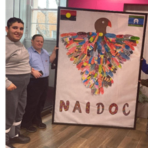 Two people stand beside a large poster featuring a bird made from colorful feathers and the word "NAIDOC," with Aboriginal and Torres Strait Islander flags at the top corners.