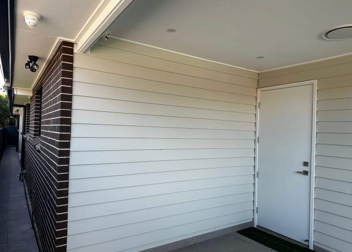 CHESTERHILL 2 1 A white door on a house with light siding and a brick corner is accompanied by a security camera and light fixture above.