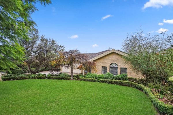 Kellyville 1 Single-story brick house with arched windows, surrounded by a neatly trimmed lawn, hedges, and several mature trees under a blue sky.