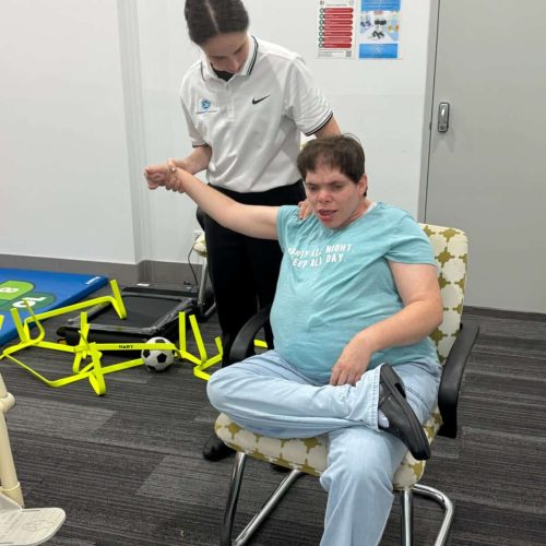 A support worker assists a seated person with mobility exercises in a therapy room equipped with sports and exercise equipment, fostering a sense of achievement with each milestone reached.
