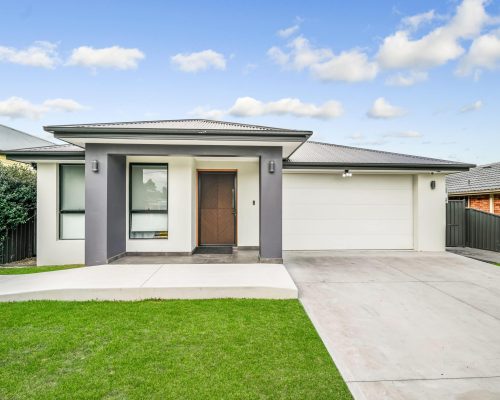 Single-story modern house with gray and white exterior, large front window, wooden door, double garage, concrete driveway, and neatly trimmed grass lawn.