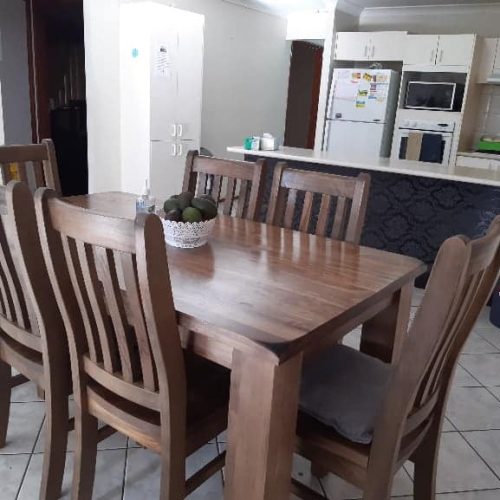 Wooden dining table with six chairs in a kitchen area. A fruit bowl is on the table. The background includes a refrigerator and cupboards.