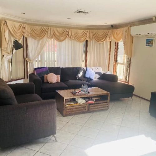 Living room with dark sofas, a wooden coffee table, and beige curtains. A floor lamp stands beside the sofa, and there are paintings on the walls. Air conditioner mounted on the wall.