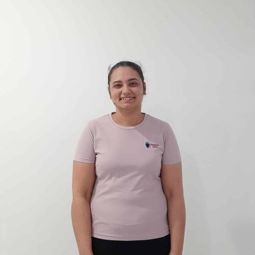 A person with tied-back hair is standing against a plain white wall, wearing a light pink t-shirt and black pants, and smiling at the camera.