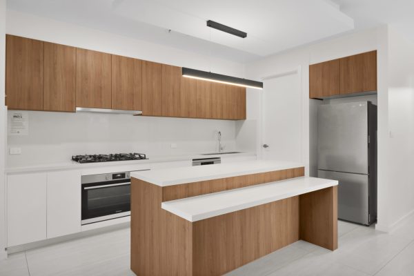 Penrith 3 3 Modern kitchen with wooden cabinets, a white countertop, built-in appliances, and a stainless steel refrigerator. A linear pendant light hangs above the kitchen island.