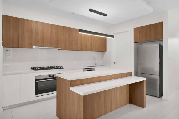 Modern kitchen with wooden cabinets, a white countertop, built-in appliances, and a stainless steel refrigerator. A linear pendant light hangs above the kitchen island.