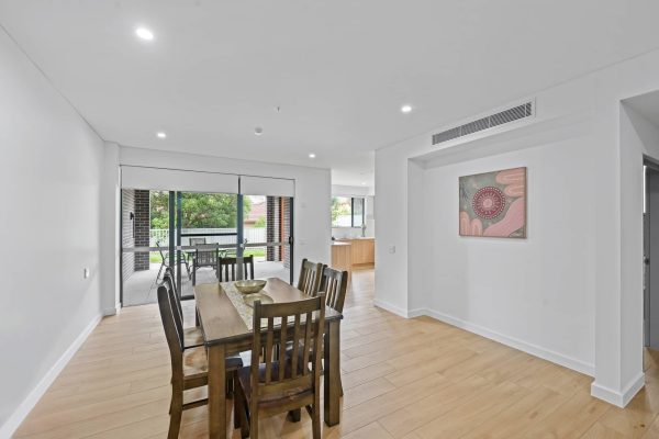 Dining area with a wooden table and six chairs on a light wood floor. Glass doors open to a patio. A kitchen is visible in the background, with a painting on the wall to the right.