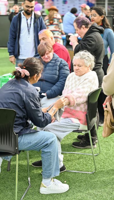 A group of people are seated outdoors on a grassy area. One woman is applying nail polish to an elderly woman's nails while others watch and wait nearby.