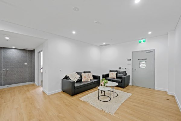 Merrylands SDA 5 Minimalist living room with two black sofas, a round coffee table on a rug, and a door labeled "Exit." Walls are white and floor is light wood. Coats of arms visible on walls.