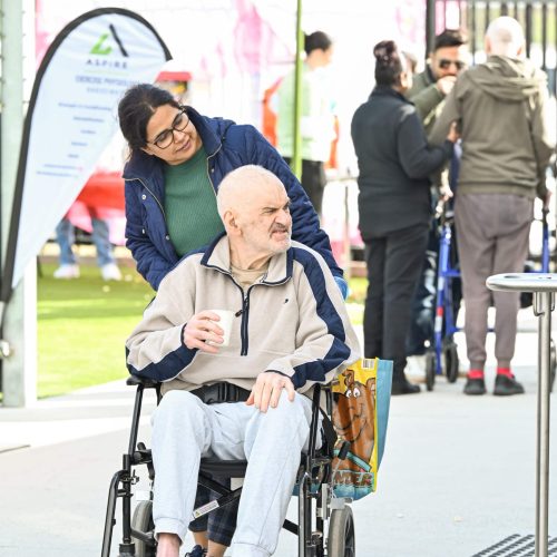 A woman pushes an elderly man in a wheelchair. He holds a drink, and there's a bag on his lap. People and a tent are visible in the background.