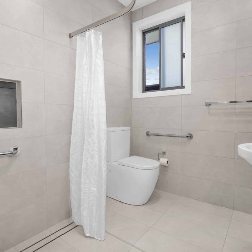 A modern, accessible bathroom with beige tiles, featuring a toilet, sink, and a shower with a white curtain. Grab bars are installed near the toilet and shower area. A window is on the wall.