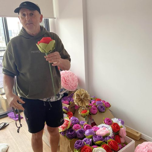 A person stands indoors holding a large artificial rose, with bags and boxes of colorful artificial flowers on the floor nearby.